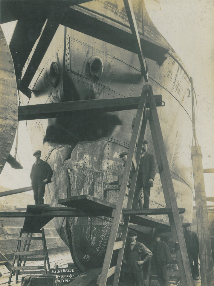 Repairs are carried out to the SS Straide's bow. A worker peers out through the large dark hole in the centre.