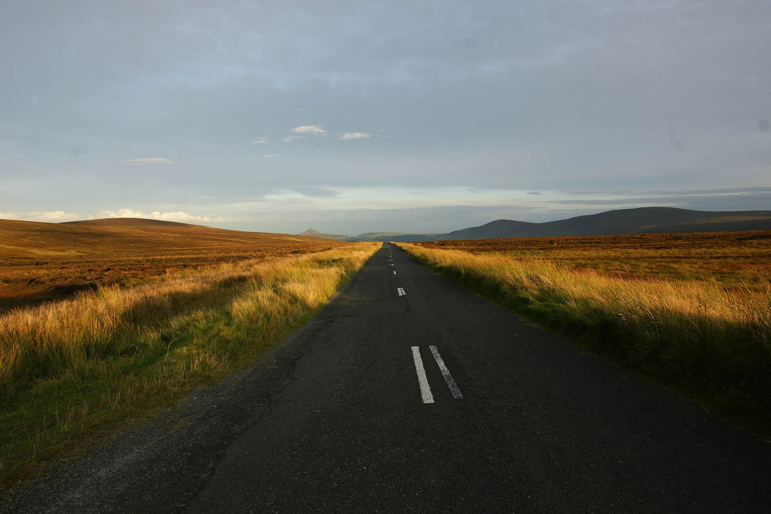 A clean empty country road flanked either side by mountain moorlands. Sunset shines through the glass.